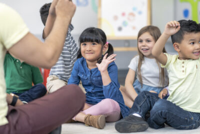 A multi-ethnic group of kindergarten students is sitting with their legs crossed on the floor in their classroom. The teacher is sitting on the floor facing the children. The happy kids are smiling and listening to the teacher. The image is focused on a little Asian girl who is raising her hand and ready to ask a question.